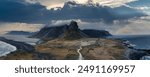 Dramatic aerial landscape of Iceland featuring a winding road through a misty valley, volcanic beach, and majestic mountain range shrouded in clouds.