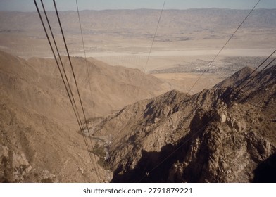Dramatic aerial perspective from a cable car looking down over a vast arid desert valley with rugged rocky mountain terrain and a distant city below.
