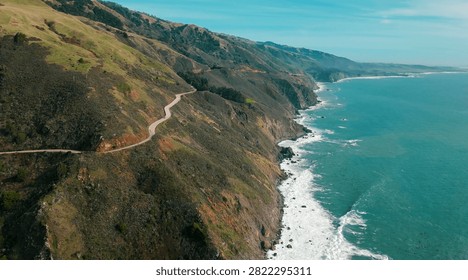 A dramatic aerial perspective captures the winding Pacific Coast Highway clinging to the rugged cliffs of Big Sur as they plunge into the turquoise waters of the Pacific Ocean. Rolling green and golden hillsides cascade down to rocky shorelines where white surf breaks against dark coastal rocks, stretching into the misty distance. Bright midday light illuminates the vivid contrast between the emerald hills, red-ochre exposed earth, and the brilliant blue-green sea below.