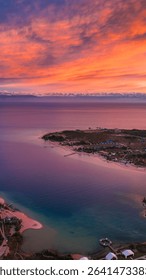 Dramatic aerial perspective of Issyk Kul Lake during a fiery orange and red sunset, highlighting the winding coastline and distant resort areas