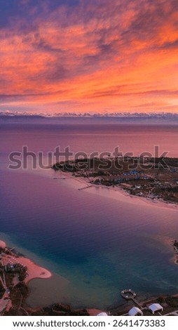 Dramatic aerial perspective of Issyk Kul Lake during a fiery orange and red sunset, highlighting the winding coastline and distant resort areas