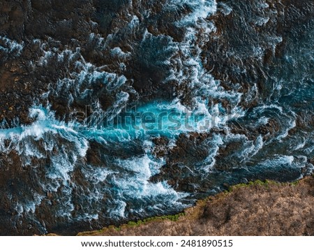 Dramatic aerial view of a bright blue river flowing through dark, rocky banks in Iceland. The water churns and froths, creating a stunning natural scene.