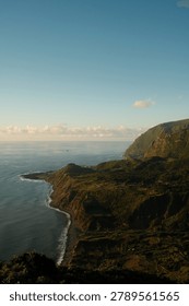 Dramatic aerial view captures rugged green coastal cliffs plunging into the Atlantic Ocean during golden hour, with terraced hillsides and a winding shoreline stretching toward the horizon. Warm low-angle sunlight illuminates volcanic rock formations and lush vegetation typical of Macaronesian island landscapes, while soft clouds gather at the distant horizon. Ideal for travel, nature, adventure, and destination marketing content.