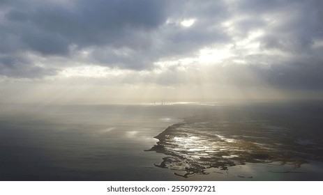 A dramatic aerial view of a coastal landscape with a cloudy sky. Sunbeams pierce through the clouds, casting a golden glow on the rugged coastline and the calm ocean waters. - Powered by Shutterstock