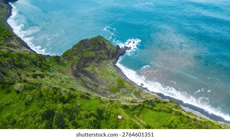 Dramatic aerial view of Madeira's lush green coastal cliffs meeting the vibrant blue Atlantic, with waves crashing on a dark pebble beach. Arco de Sao Jorge