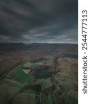 Dramatic aerial view of a moody landscape under dark clouds, featuring rolling hills, farmland, and winding country roads.