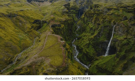 Dramatic aerial view of a moss covered canyon in Iceland, featuring steep cliffs, a winding river, two cascading waterfalls, and a narrow ridge trail.