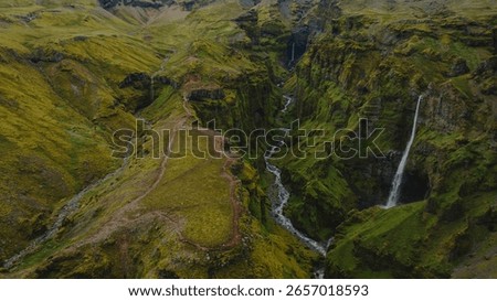 Dramatic aerial view of a moss covered canyon in Iceland, featuring steep cliffs, a winding river, two cascading waterfalls, and a narrow ridge trail.