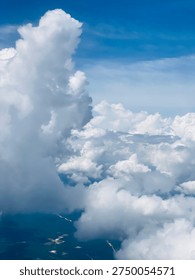 Dramatic aerial view of towering white clouds rising above the landscape, photographed from an airplane window at high altitude. The massive cloud formations create a majestic sky scene while glimpses of land appear far below through the cloud gaps.