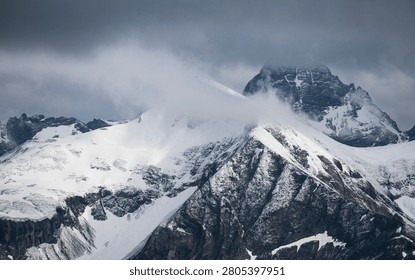 A dramatic alpine landscape featuring a rugged, snow-covered mountain ridge under a moody sky. The sharp crest of the mountain extends diagonally through the frame, with steep rocky slopes partially covered in fresh snow. Thick clouds drift across the peaks, obscuring parts of the background mountains and creating an atmosphere of mystery and grandeur. The contrast between the dark rock faces, bright white snowfields, and stormy gray clouds emphasizes the harsh beauty of the high-altitude environment. The image conveys a sense of isolation, power, and the untamed nature of the alpine wilderness.
Taken in the Allgäuer Alps on the Nebelhorn.