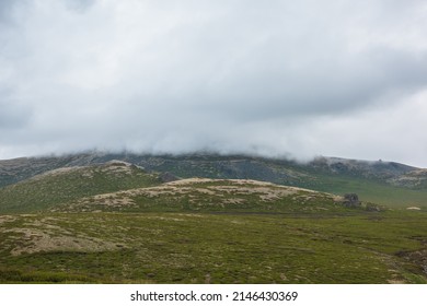 Dramatic alpine landscape with hills and mountains in gray low clouds. Bleak atmospheric scenery of tundra under lead gray sky. Gloomy minimalist view to green hills among low rainy clouds in overcast
