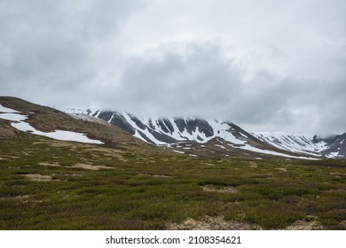 Dramatic alpine landscape with snowy mountains in gray low clouds. Bleak atmospheric scenery of tundra under lead gray sky. Gloomy minimalist view to mountain range among low rainy clouds in overcast. - Shutterstock ID 2108354621