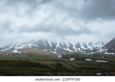 Dramatic alpine landscape with snowy mountains in gray low clouds. Bleak atmospheric scenery of tundra under lead gray sky. Gloomy minimalist view to mountain range among low rainy clouds in overcast.