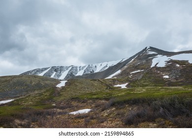 Dramatic alpine landscape with snowy mountains under gray clouds. Bleak overcast scenery with mountain range under rainy clouds. Gloomy atmospheric view to pyramidal mountain top under lead gray sky.