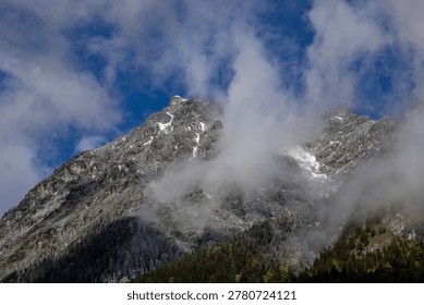 Dramatic alpine mountain peaks covered with traces of late spring snow, wrapped in low moving clouds beneath a vivid blue sky in the Bavarian Alps.