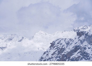 A dramatic alpine scenery showcasing jagged, dark rocky mountain slopes contrasted against snow-covered background peaks under an overcast winter sky.