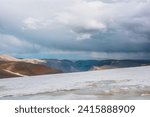 Dramatic alpine top view from flat sloping glacier on precipice edge to deep gorge between colorful sunlit cliffs and mountain range silhouette in rain under lead gray cloudy sky. Rainy grey clouds.