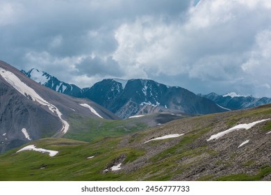 Dramatic alpine view to big rocky ridge and stony green hills under rainy gray sky. Overcast landscape with large wide snow-capped mountain range. Beautiful hilly terrain and snowy mountains far away. - Powered by Shutterstock