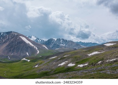 Dramatic alpine view to big rocky ridge and stony green hills under rainy gray sky. Overcast landscape with large wide snow-capped mountain range. Beautiful hilly terrain and snowy mountains far away. - Powered by Shutterstock