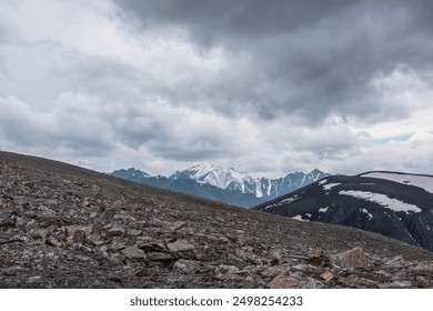 Dramatic alpine view from high stony pass to big snowy mountain top under gray cloudy sky. Large snow castle in gloomy rainy weather. Cold atmospheric landscape with sharp stones in wild mountains.