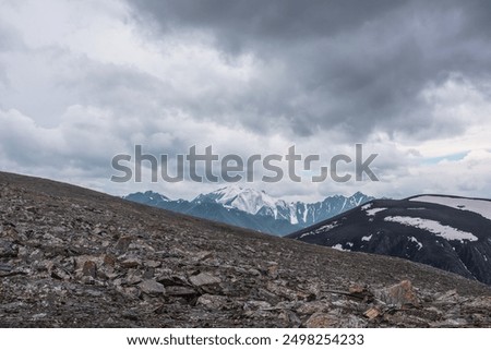 Dramatic alpine view from high stony pass to big snowy mountain top under gray cloudy sky. Large snow castle in gloomy rainy weather. Cold atmospheric landscape with sharp stones in wild mountains.