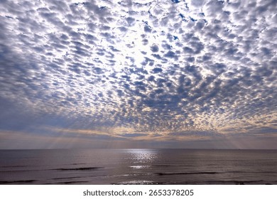 Dramatic altocumulus cloudscape over calm sea, sunrays breaking through the textured sky and shimmering on the water's surface at twilight.