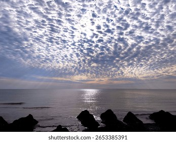 Dramatic altocumulus cloudscape over calm sea, sunrays breaking through the textured sky and shimmering on the water's surface at twilight.