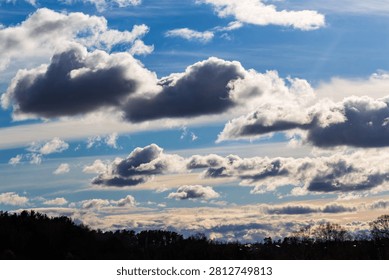 Dramatic April sky with fluffy white and dark grey clouds drifting over a dark forest silhouette, capturing a crisp spring afternoon mood with dynamic light and soft celestial textures