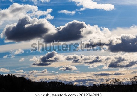 Dramatic April sky with fluffy white and dark grey clouds drifting over a dark forest silhouette, capturing a crisp spring afternoon mood with dynamic light and soft celestial textures