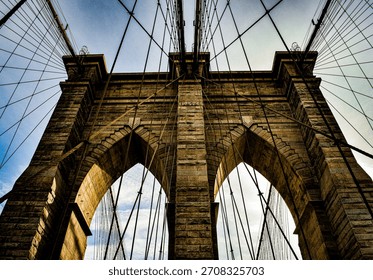 Dramatic architectural view of the Brooklyn Bridge, captured from below with strong lines, stone arches and converging cables creating a powerful geometric composition.