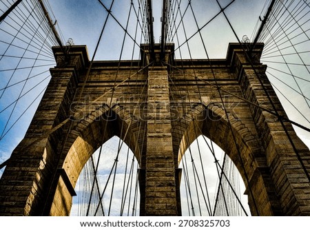Dramatic architectural view of the Brooklyn Bridge, captured from below with strong lines, stone arches and converging cables creating a powerful geometric composition.