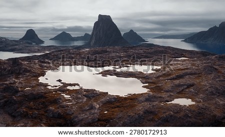 Dramatic arctic landscape featuring rocky, dark terrain with small pools of water and sharp, majestic mountain peaks in the background