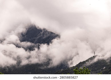 A dramatic, atmospheric capture of jagged mountain peaks emerging from thick, rolling clouds and mist. The rugged, forest-covered slopes are partially obscured by the ethereal fog, emphasizing the sheer scale and mystical quality of the highland terrain. An electrical transmission tower stands prominently on a ridge, adding a touch of modern infrastructure to the wild, untouched landscape.