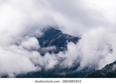 A dramatic, atmospheric capture of jagged mountain peaks emerging from thick, rolling clouds and mist. The rugged, forest-covered slopes are partially obscured by the ethereal fog, emphasizing the sheer scale and mystical quality of the highland terrain. An electrical transmission tower stands prominently on a ridge, adding a touch of modern infrastructure to the wild, untouched landscape.