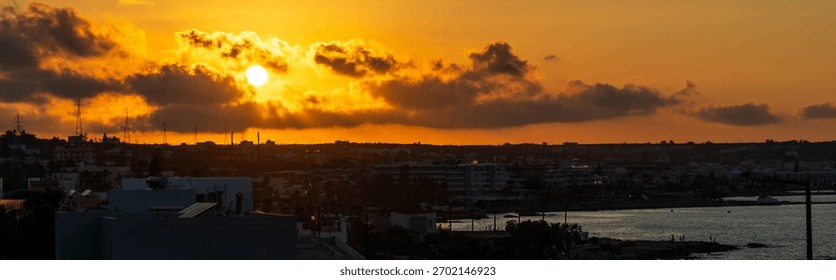 Dramatic August sunset over coastal Crete with warm orange light, clouds illuminated by the low sun and town buildings silhouetted against the colorful evening sky.