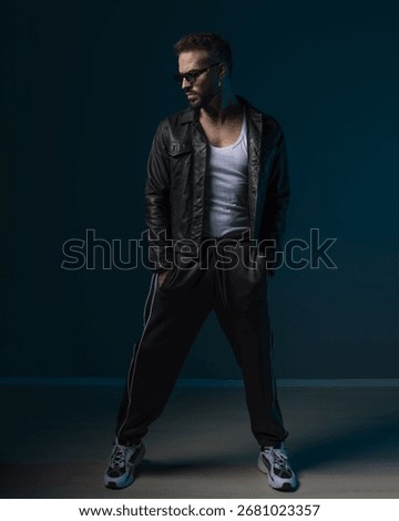 dramatic bad boy with sunglasses looking to side and posing with hands in pockets in front of blue background