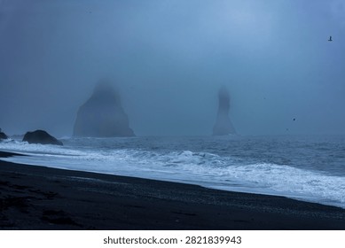 Dramatic basalt sea stacks emerge from fog-shrouded waters off Iceland's black sand beach, with rolling white waves and moody blue twilight