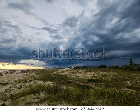 A dramatic beach scene with a stormy sky, shrouded in dark, menacing clouds, contrasting with the golden light of the sunset in the background.