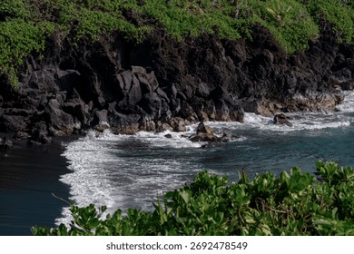 Dramatic black sand beach waves with volcanic rocks and lush green foliage at Wai'anapanapa State Park along the Road to Hana in Maui, Hawaii, USA