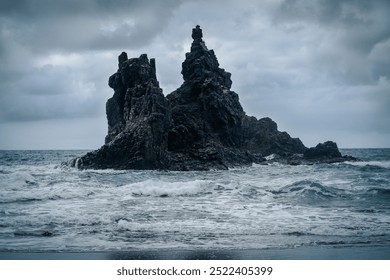 Dramatic black volcanic basalt formations and rocks amidst stormy ocean in cloudy weather.