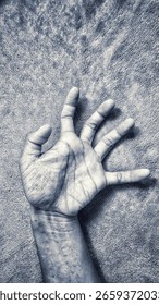 A dramatic black and white close-up captures a human hand with fingers splayed, palm facing slightly upwards, resting on a textured surface. The monochrome treatment emphasizes the intricate lines