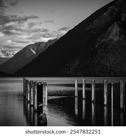 A dramatic black and white composition featuring a tranquil dock, serene waters, and towering mountains in the background. A striking blend of light, shadow, and peaceful scenery. - Powered by Shutterstock