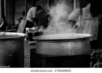 A dramatic black and white image of large, hammered metal pots steaming over a fire, with chefs working in the background at an Indian street food stall in Ahmedabad railway station