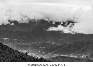 A dramatic black and white landscape capturing a vast, remote mountain valley. The monochrome presentation emphasizes the rich textures of the dense evergreen forests and the striking contrast of the heavy, low-hanging clouds rolling over the mountain peaks. Down in the valley, distant settlements and winding roads highlight the immense scale of the surrounding wilderness. This moody, atmospheric shot perfectly conveys the rugged beauty and quiet isolation of the natural world.