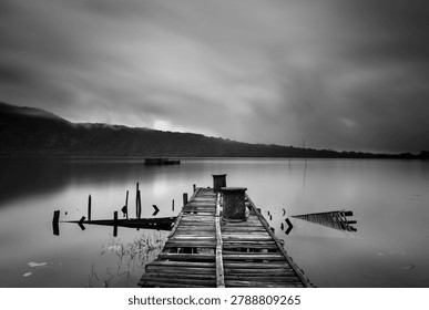Dramatic black and white long exposure landscape of a broken wooden pier on a calm mountain lake under an overcast sky
