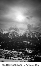 Dramatic black and white panoramic view of snow-covered alpine mountains towering above a small village in a deep valley. Low clouds and mist surround the rugged peaks, creating a moody and atmospheric winter landscape. Perfect for themes such as mountain travel, wilderness, dramatic weather, scenic alpine photography, and monochrome nature art.