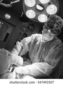 Dramatic Black And White Photo Of The Operating Surgeon In The Surgery Room. Surgeon In Mask And Uniform And Surgery Light Beside