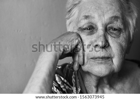 Dramatic black and white portrait of an elderly woman with gray hair. She looks sadly at the camera and rests her head on her hand. Face close-up.