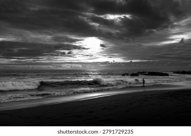 Dramatic black and white seascape featuring rolling waves, dark storm clouds, and a solitary figure standing near the shoreline. The contrast between the turbulent sky and reflective beach creates a powerful and atmospheric coastal landscape, evoking themes of solitude, contemplation, and the raw beauty of nature.