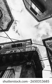 A dramatic black and white upward view of old Sicilian buildings with balconies, wires, and cloudy sky.